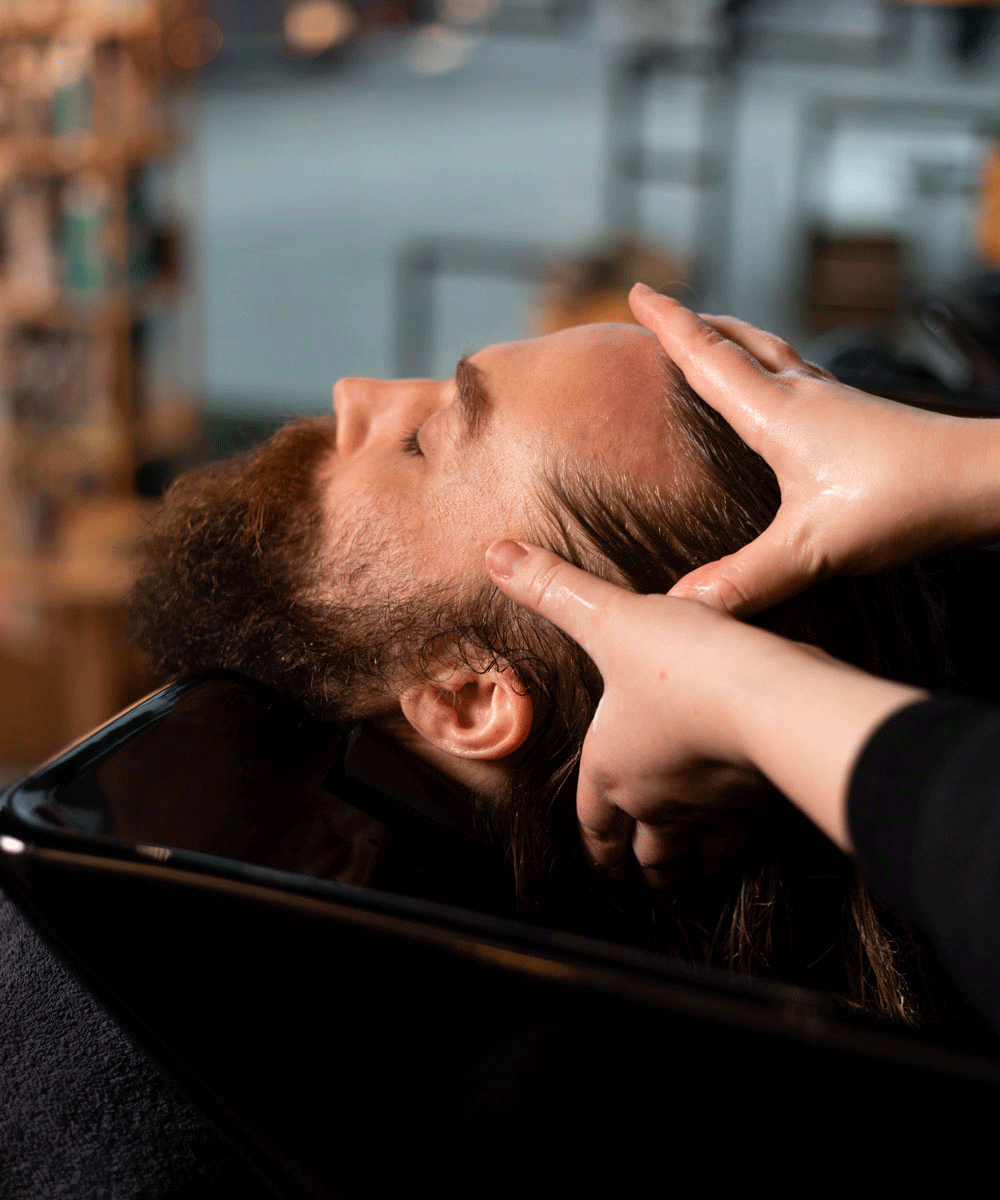 Mann med langt hår får avslappende hodebunnsmassasje under hårvask hos Nikita Hair.