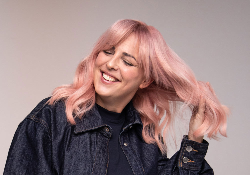 Person med pudderrosa hår i myke bølger, fotografert i bevegelse mot nøytral bakgrunn