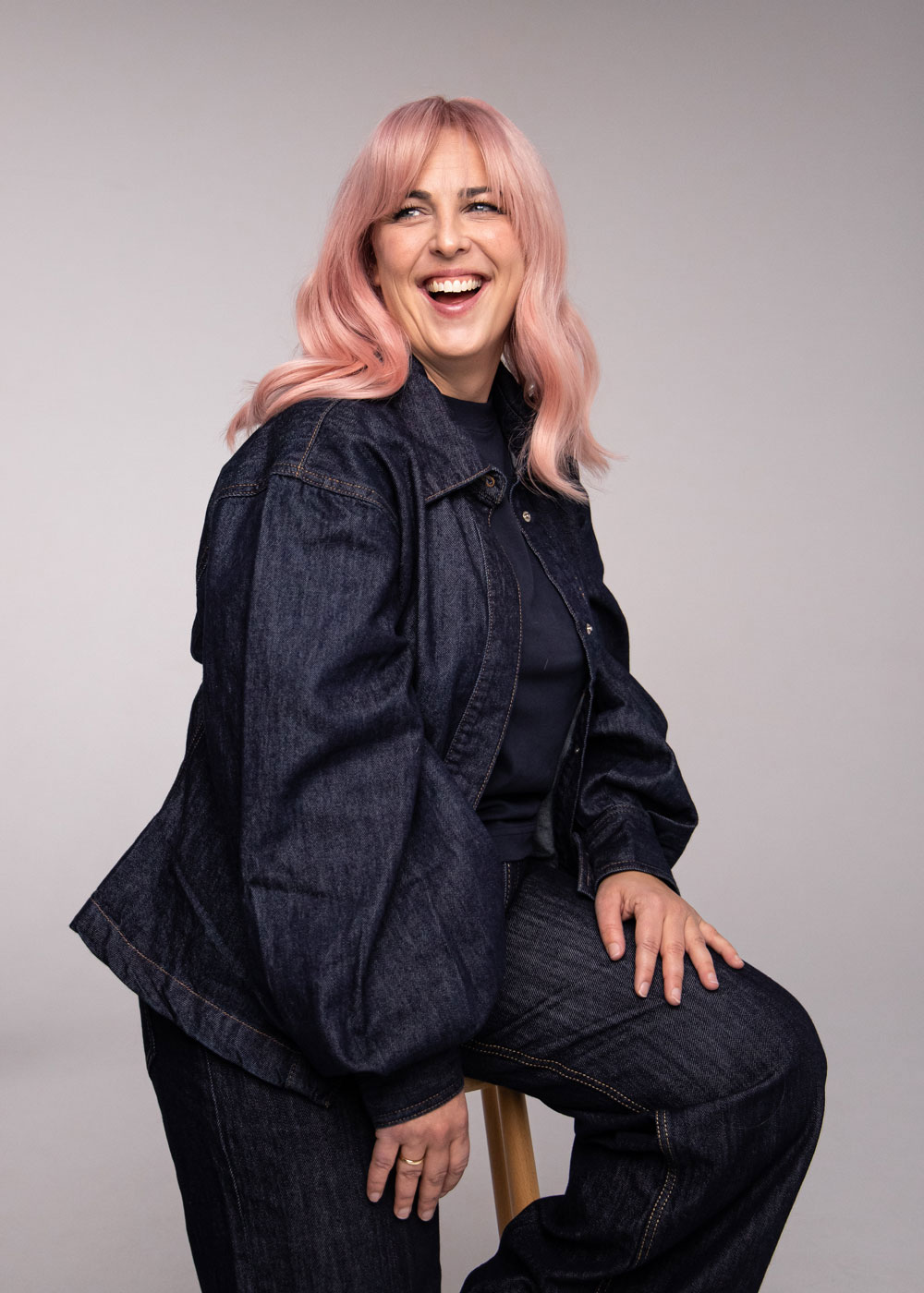 Person med pudderrosa hår i myke lengder, sittende i denimantrekk mot nøytral bakgrunn